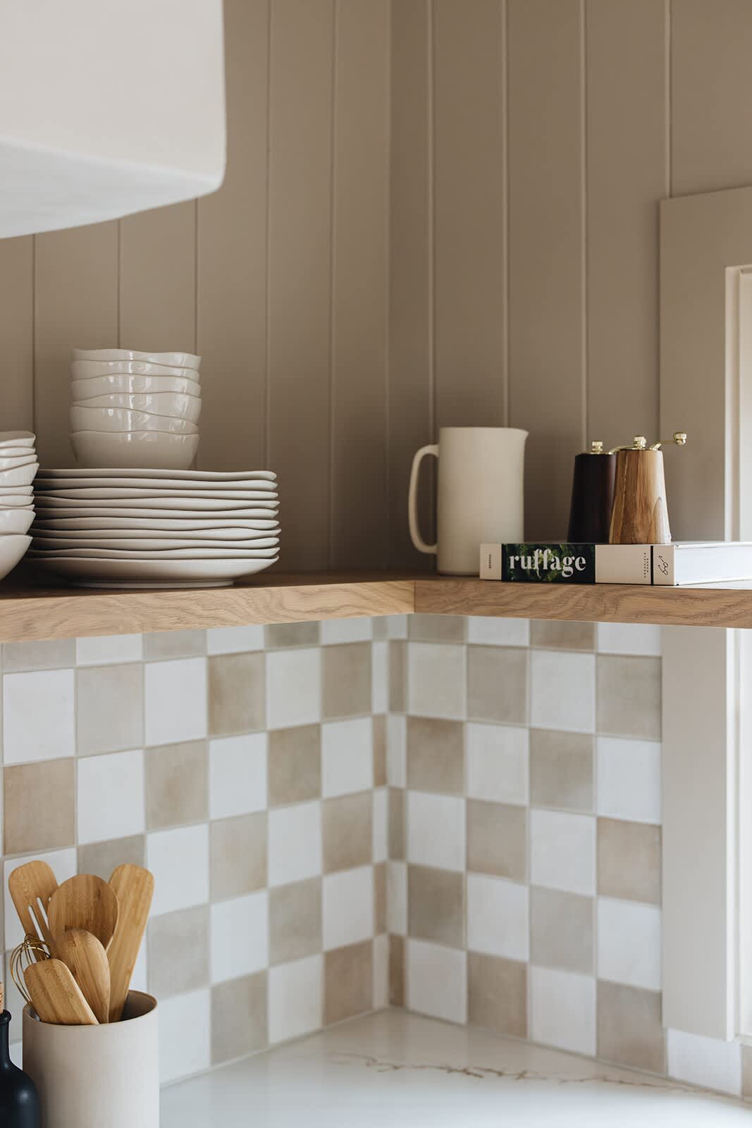 Corner view of the kitchen with stacked white plates and bowls on a floating wooden shelf. The wall features vertical wood paneling and a checkered tile backsplash, with wooden utensils in a holder on the counter.