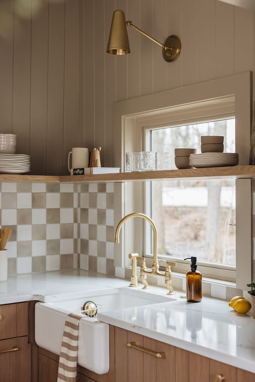 Detailed view of the kitchen sink area featuring a white farmhouse sink, a prominent brass faucet, and a checkered tile backsplash. A window overlooks a snowy outdoor scene, and a brass wall sconce illuminates the space.