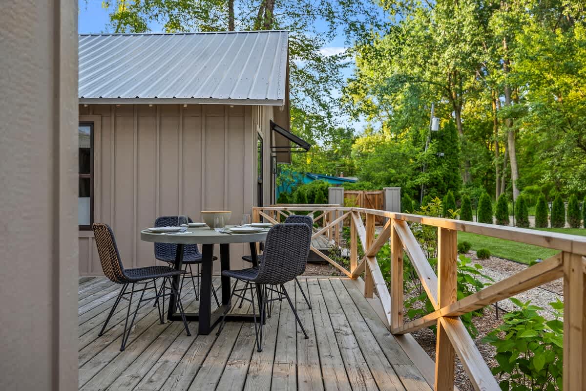 A close-up of a wooden deck with a round outdoor dining table and four woven chairs, set against a backdrop of lush green trees. The deck features light wooden railings and is surrounded by small, well-maintained plants.