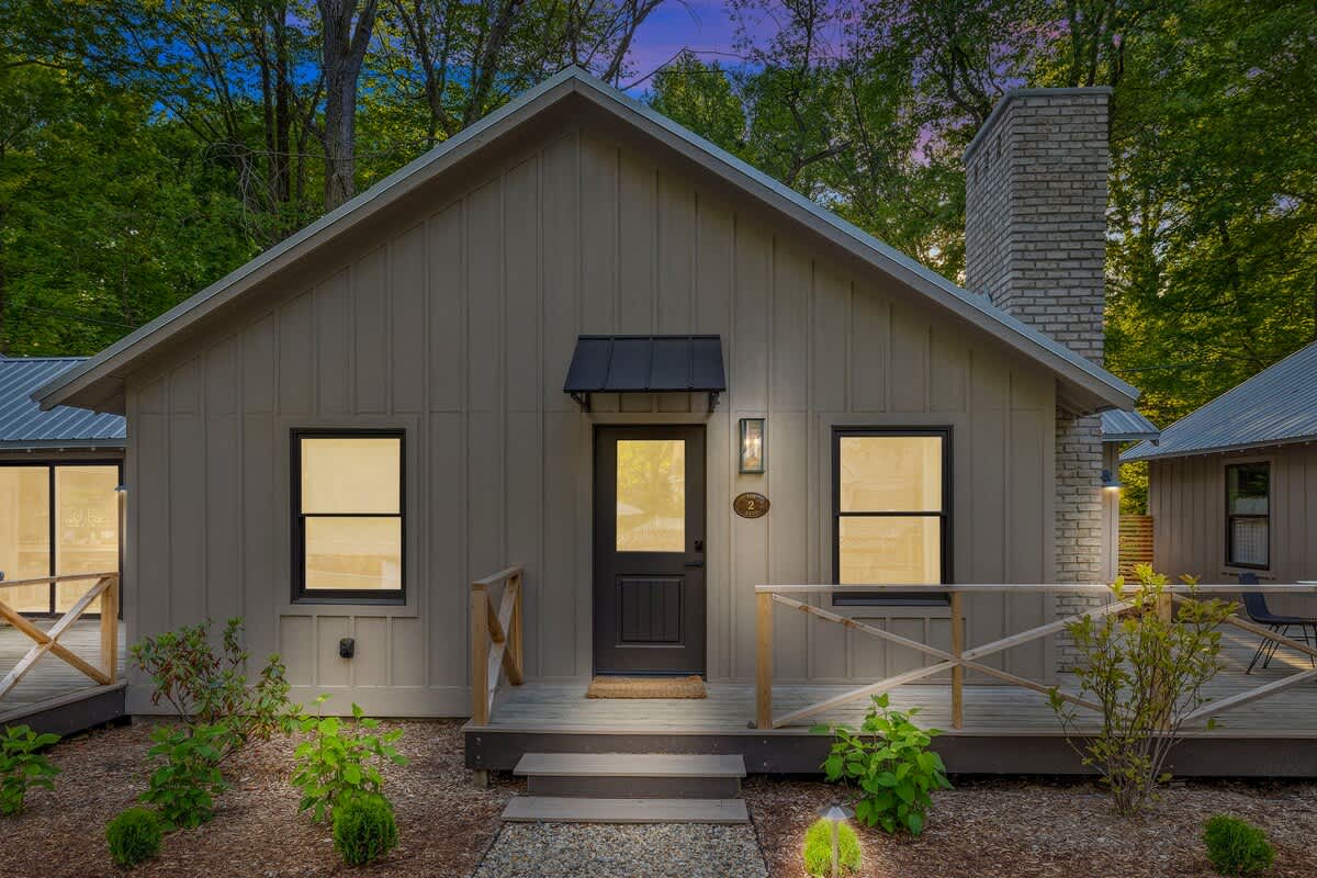 The front exterior of a modern cabin features light beige vertical siding, a dark front door with a small black awning, and two large windows with black frames. A small wooden deck with railings and steps leads to the entrance, surrounded by lush greenery.