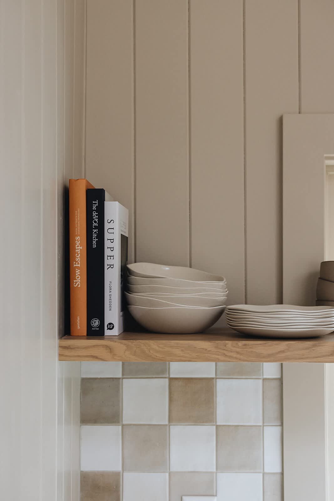 A close-up of an open wooden shelf in the kitchen, holding cookbooks and stacks of neutral-toned ceramic bowls and plates, above a checkered tile backsplash.