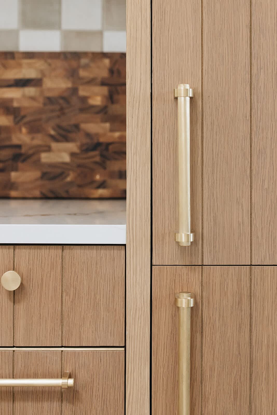 Close-up of light wood kitchen cabinetry with vertical paneling and modern brass hardware, featuring a round knob and cylindrical pulls. A butcher block cutting board is visible in the background.