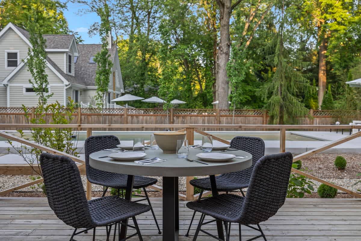 Spacious wooden deck with a round outdoor dining table and four chairs, offering a clear view of a large outdoor pool and surrounding lounge chairs, framed by mature trees.