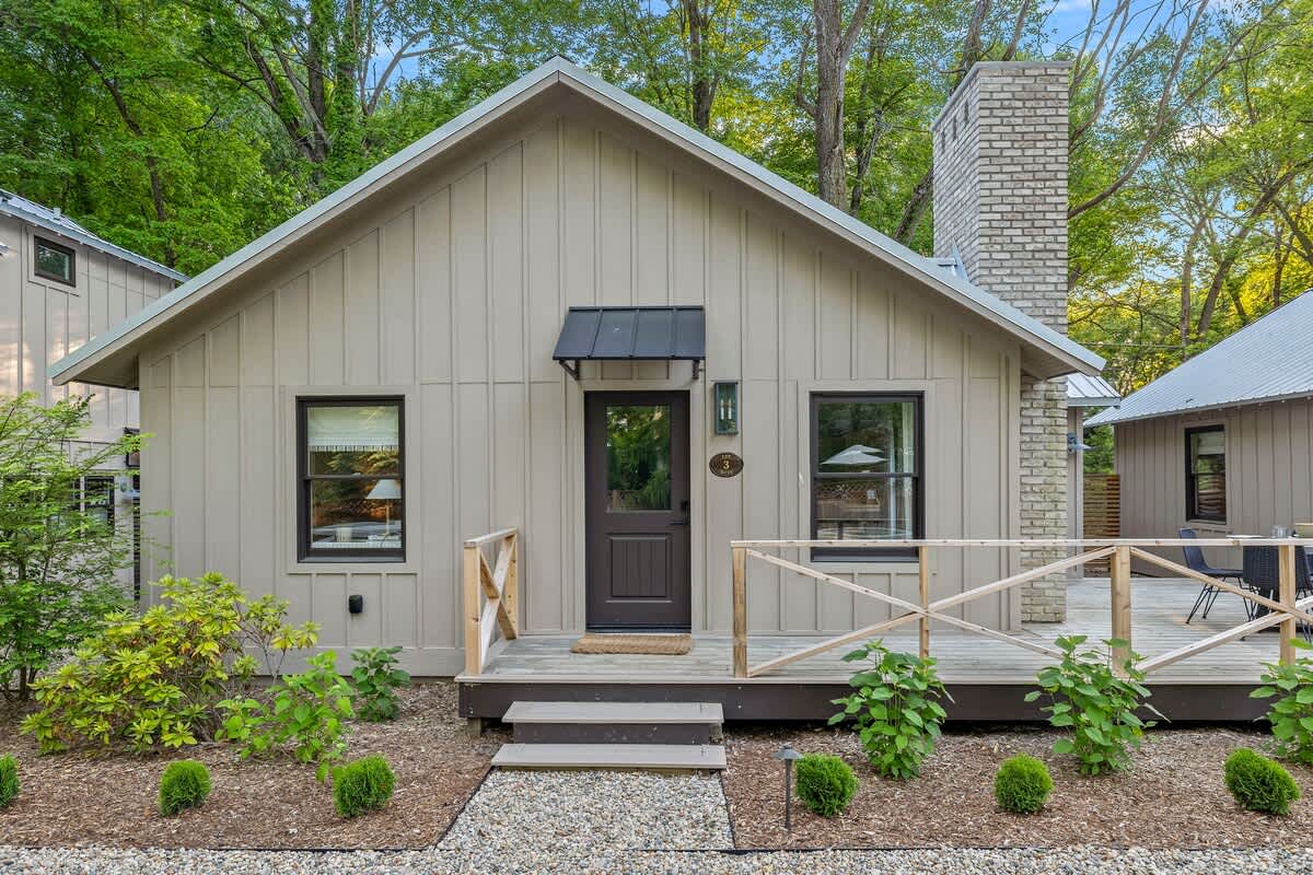 Charming cottage with light beige board-and-batten siding, a dark front door with a small awning, black-framed windows, and a small wooden deck with a railing, surrounded by landscaping.