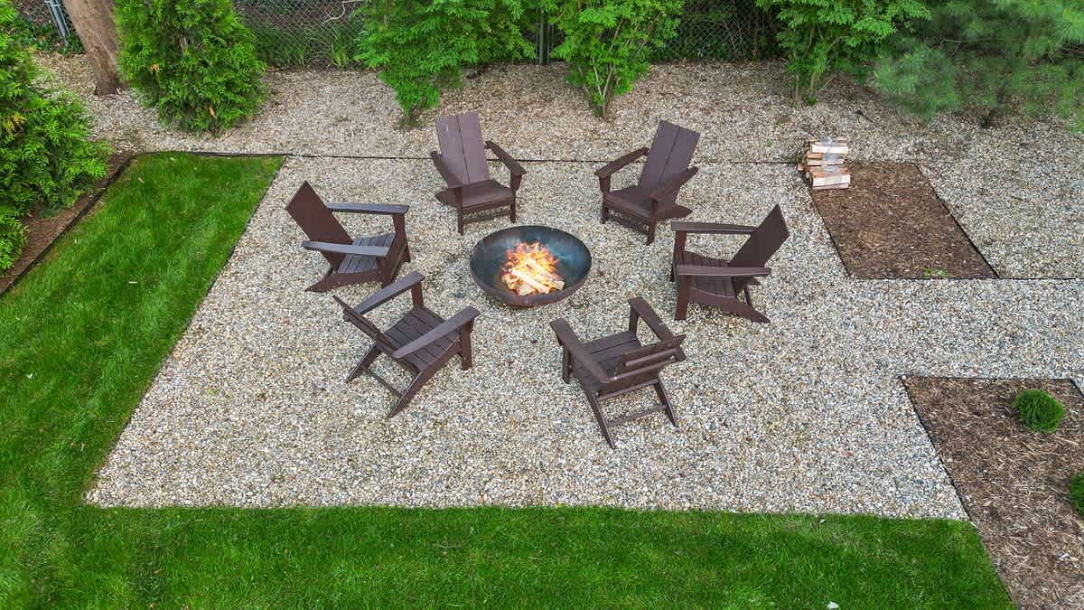 Overhead view of a circular fire pit with a burning fire, surrounded by six dark brown Adirondack chairs on a light gravel patio, bordered by a neatly trimmed green lawn and mature trees.