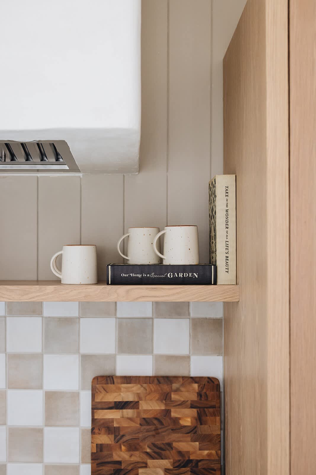 A close-up of an open wooden shelf in the kitchen, displaying white ceramic mugs and a stack of books, set against a vertical plank wall and checkered tile backsplash.