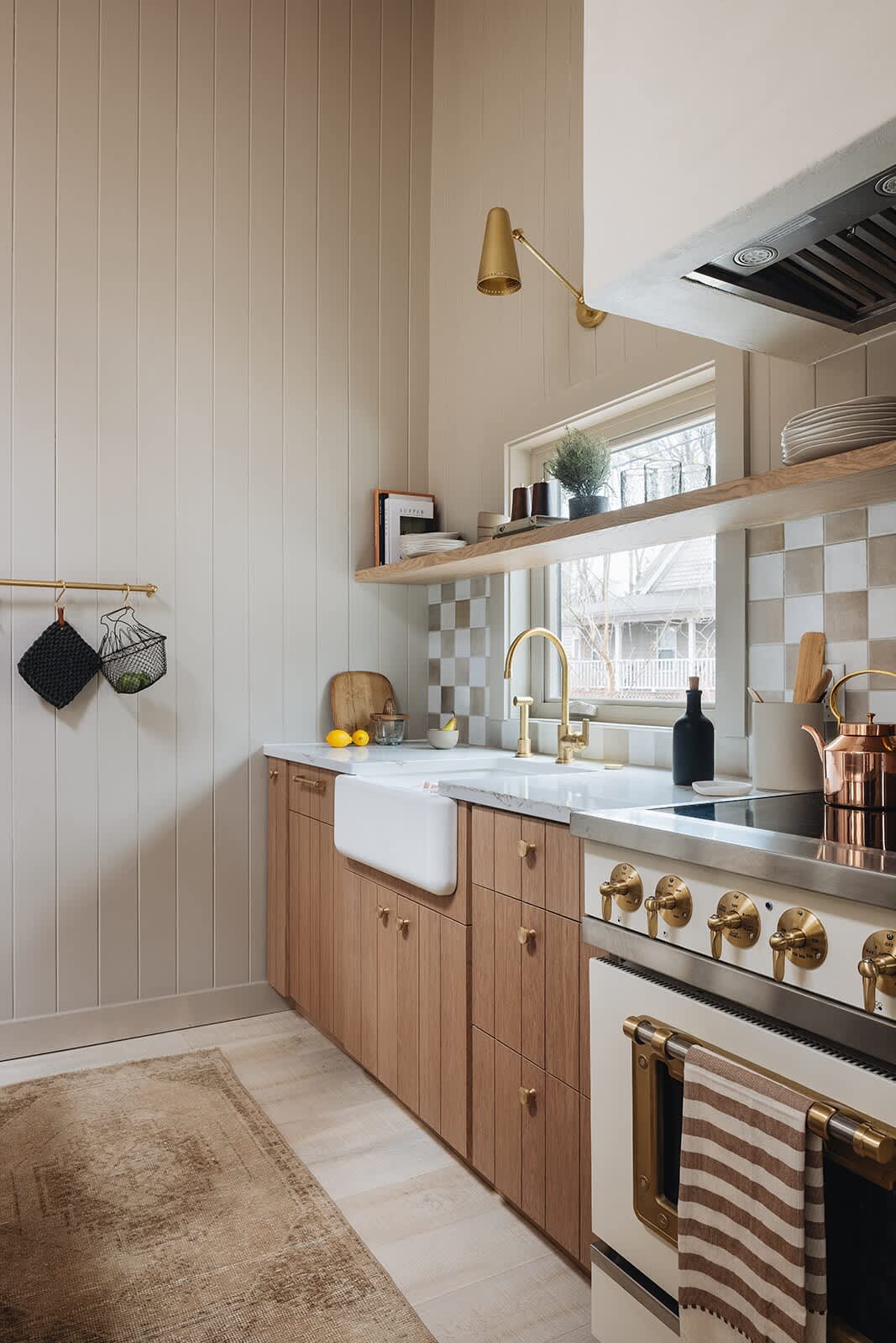 A perspective shot of the kitchen counter, featuring a white farmhouse sink, brass faucet, light wood cabinetry, and a checkered tile backsplash, with a brass wall sconce above.