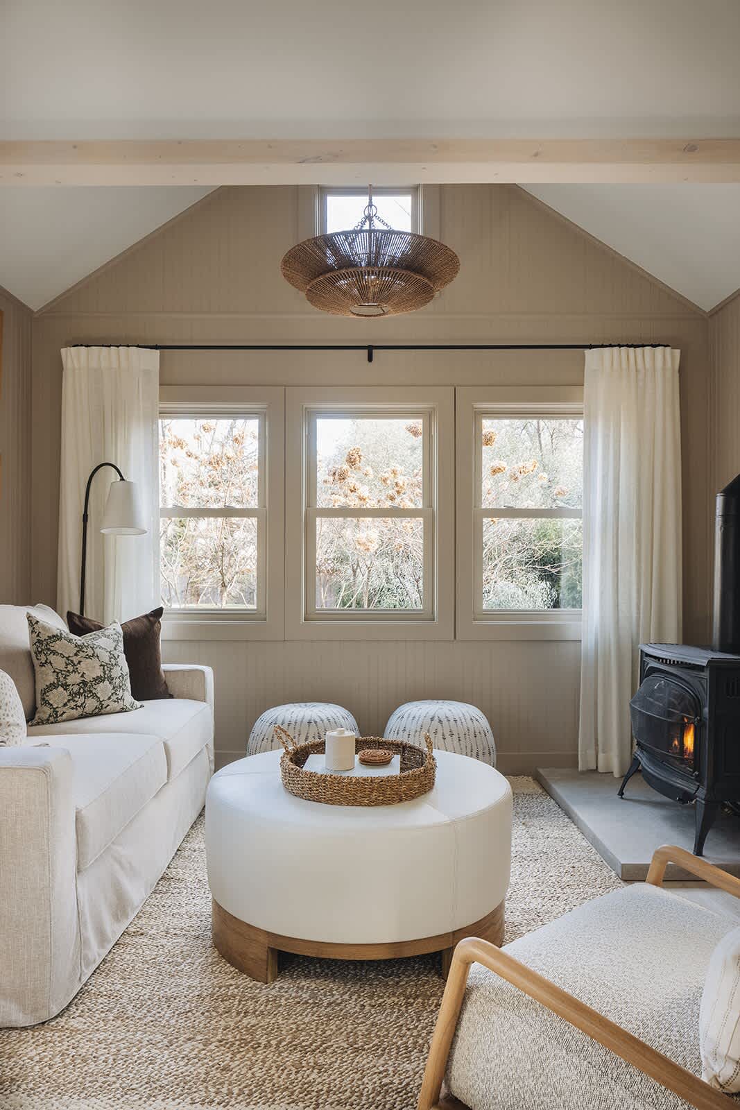 Cozy living room with a neutral color palette, featuring a white slipcovered sofa, a round white ottoman coffee table, and a black wood-burning stove, all under a vaulted ceiling with exposed beams.