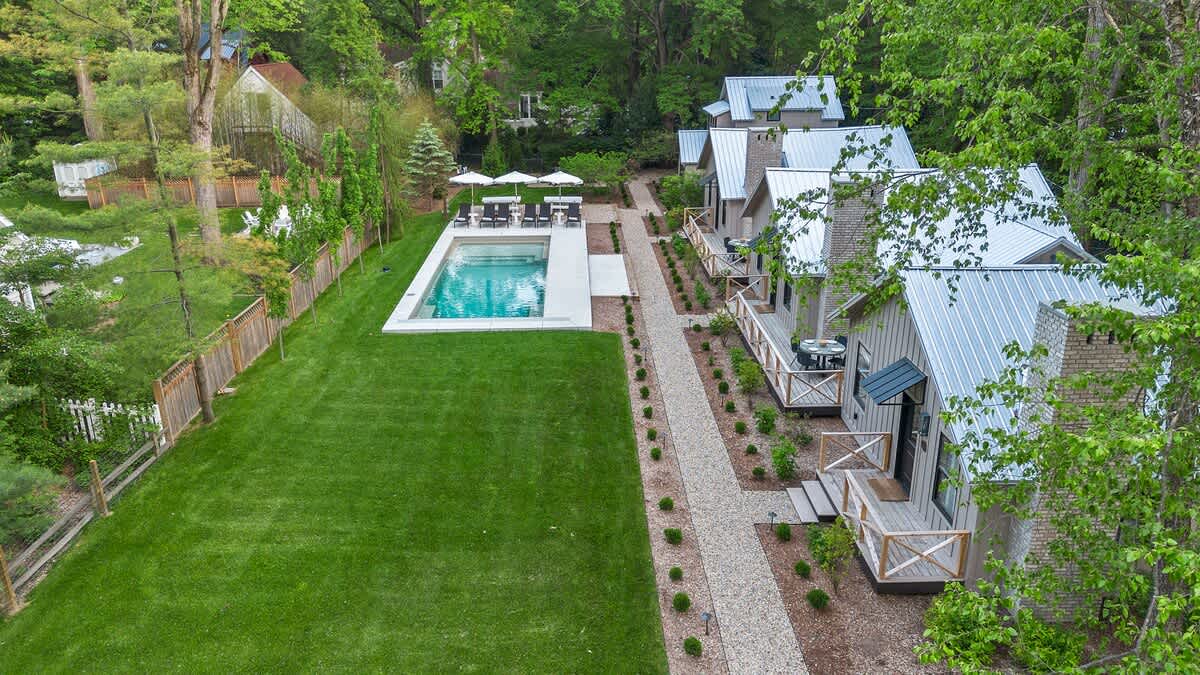 Elevated aerial view of the property, featuring a row of modern cottages with light siding and metal roofs, a long gravel pathway, a rectangular swimming pool with a surrounding patio, and a large, well-maintained green lawn, all surrounded by lush trees.