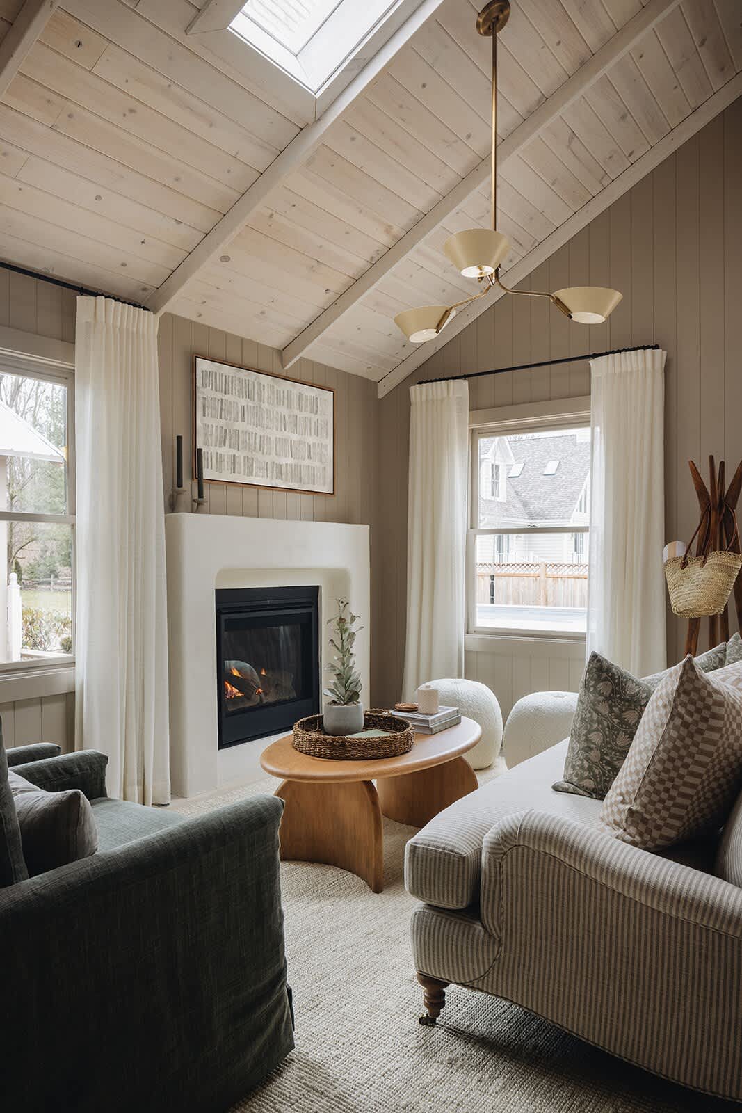A warm living room with a modern white fireplace and a lit fire, flanked by two windows with white curtains. A striped sofa and green armchairs are arranged around a wooden coffee table, under a vaulted ceiling with a brass chandelier.