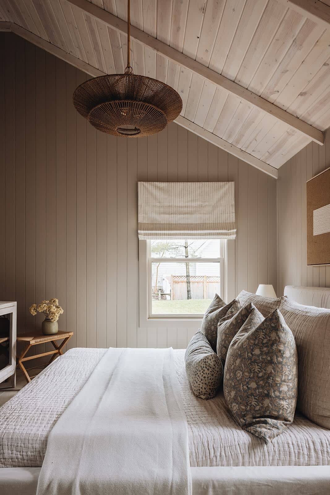 Bright bedroom with a large bed dressed in neutral linens and decorative pillows, facing a window with a striped Roman blind. The room features light beige vertical paneled walls, a vaulted white wood plank ceiling, and a woven pendant light.