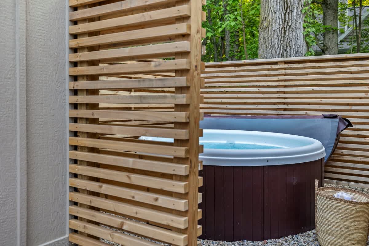 A dark brown hot tub with light blue water is partially visible behind a slatted wooden privacy screen. A woven basket sits on the gravel ground next to the tub, with lush green trees in the background.