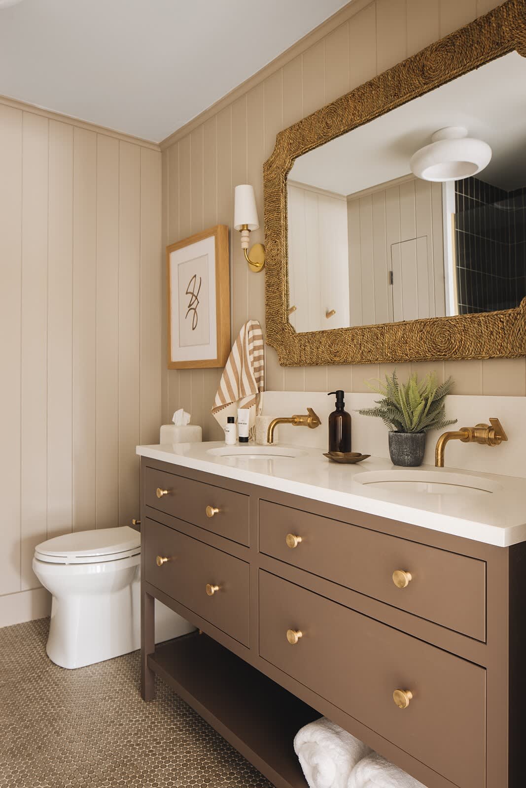 A modern bathroom featuring a double vanity with a brown cabinet, white countertop, and brass hardware. The walls are light beige vertical paneling, and a large ornate gold mirror hangs above the sinks.