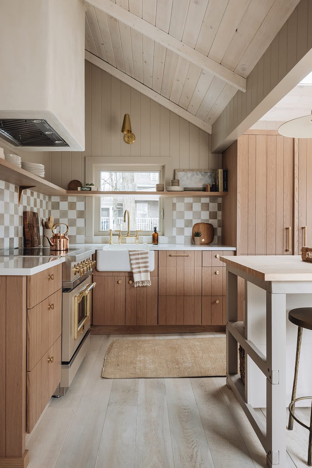 A full view of a modern kitchen with light wood cabinetry, white countertops, and a checkered tile backsplash. A beige and brass range is centered, with a white farmhouse sink under a window, and a large kitchen island with bar stools to the right.