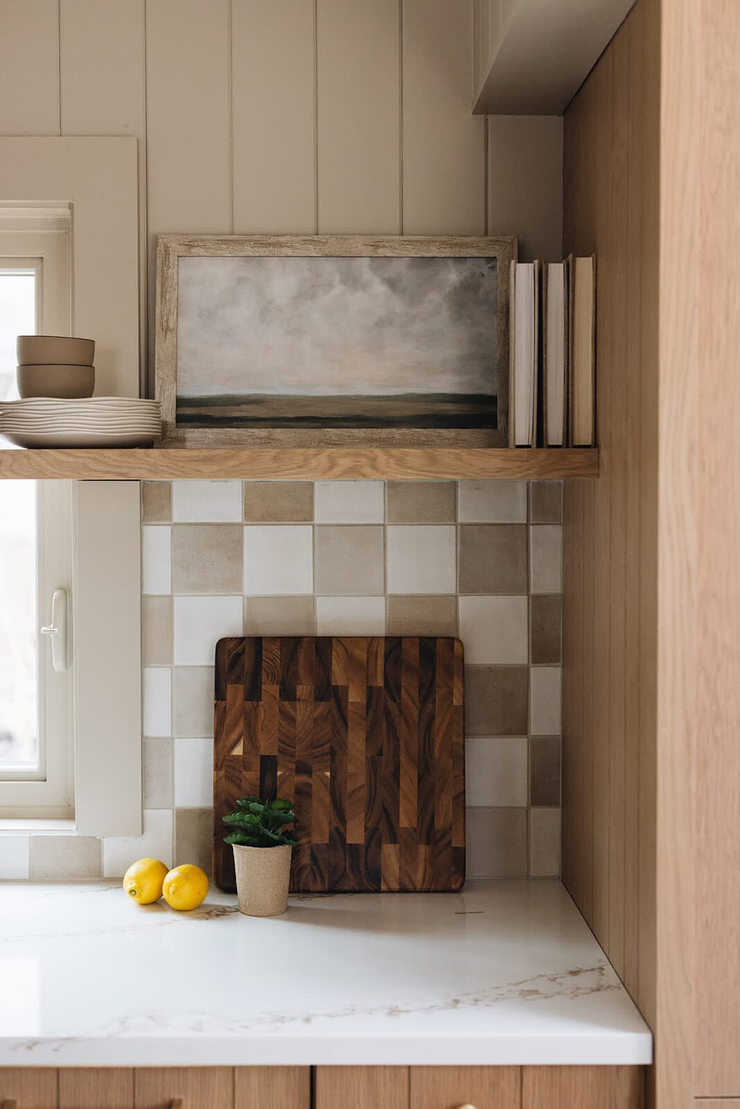 A detailed view of a kitchen counter with a checkered tile backsplash, light wood cabinets, and a white marble-look countertop, adorned with a framed landscape painting and lemons.