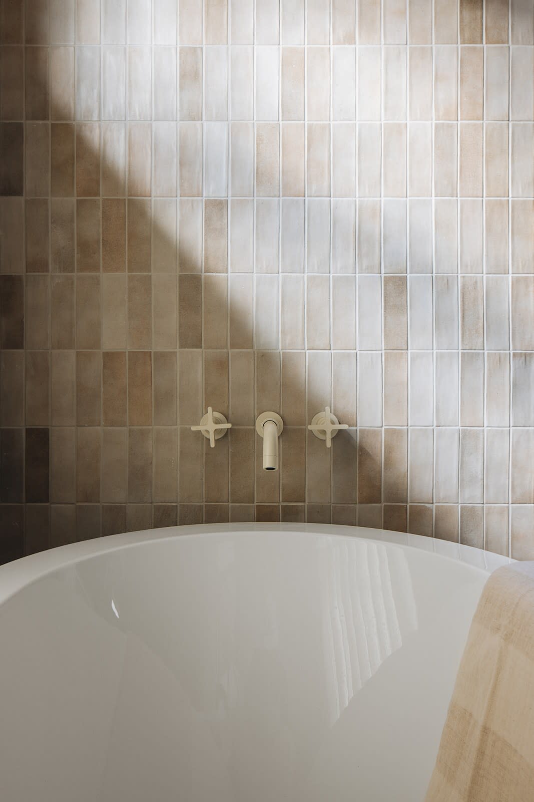 Close-up of a white freestanding bathtub against a tiled wall with varying shades of beige rectangular tiles, illuminated by natural light. A wall-mounted faucet with cross handles is visible above the tub.