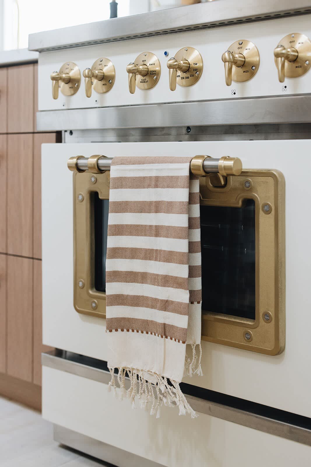 A close-up of the vintage-style oven's brass controls and handle, showing intricate detailing and a striped towel draped over the handle.