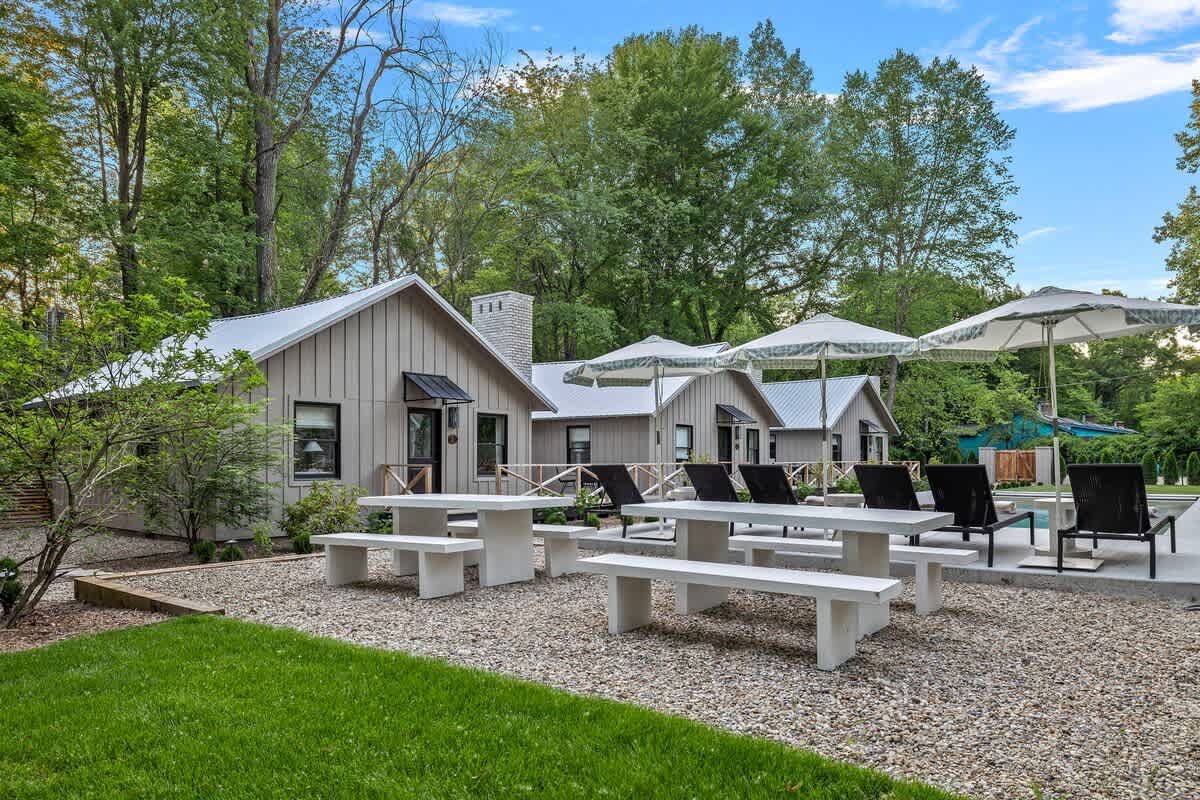 A spacious outdoor area featuring multiple light-colored cottages with dark roofs, several white picnic tables with benches on a gravel patio, and a lush green lawn, all surrounded by mature trees under a bright sky.