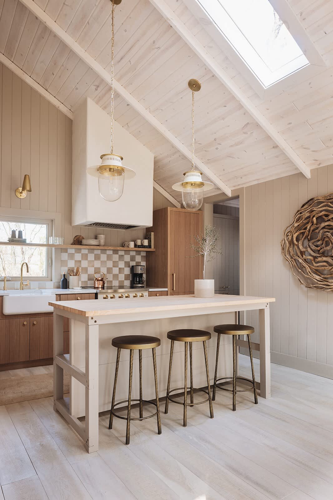 Modern kitchen with light wood cabinetry, a large white farmhouse sink, and a checkered tile backsplash, featuring a central island with three bar stools. The room boasts a vaulted whitewashed wood ceiling with pendant lights and skylights.