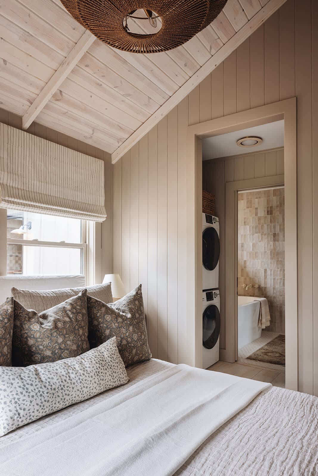 View from a bedroom with a large bed and neutral bedding, showing an open doorway leading to a laundry area with stacked washer and dryer units. The bedroom features light beige paneled walls and a vaulted white wood plank ceiling.
