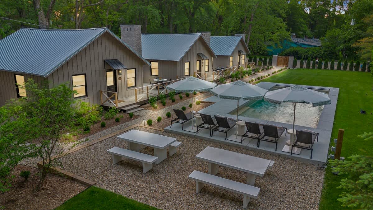 Aerial view of several modern cottages with light gray siding and metal roofs, arranged around a central gravel path, with a rectangular swimming pool, lounge chairs, and umbrellas on one side, and two concrete picnic tables on a gravel patio.
