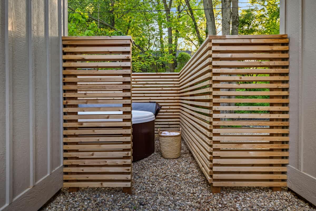 A private outdoor hot tub area enclosed by modern slatted wooden screens, set on a gravel base. A woven basket with towels is placed next to the hot tub, with lush greenery visible beyond the screens.