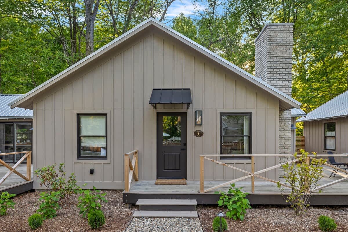A modern cottage with light beige board-and-batten siding, a dark brown front door with a black awning, and black-framed windows. A small wooden deck with steps leads to the entrance, surrounded by lush green landscaping and trees.