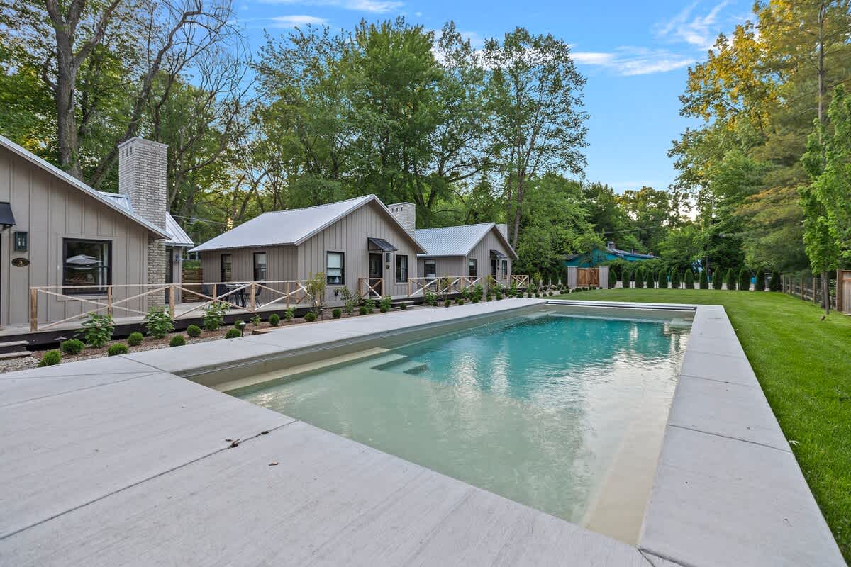 A rectangular outdoor swimming pool with light blue water, surrounded by a concrete deck, with a row of modern light-colored cottages featuring dark roofs and chimneys in the background, all set amidst lush green trees and a lawn.