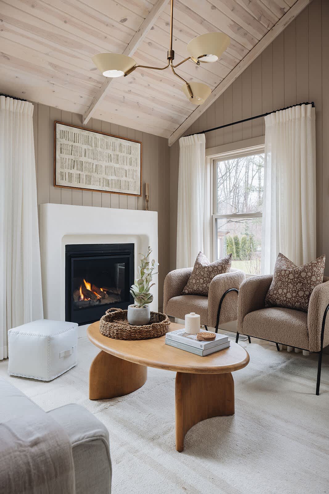 Inviting living room with a white sofa, two textured brown armchairs, and a modern wooden coffee table on a light rug, all centered around a white fireplace. The room features light wood floors and a vaulted whitewashed wood ceiling.