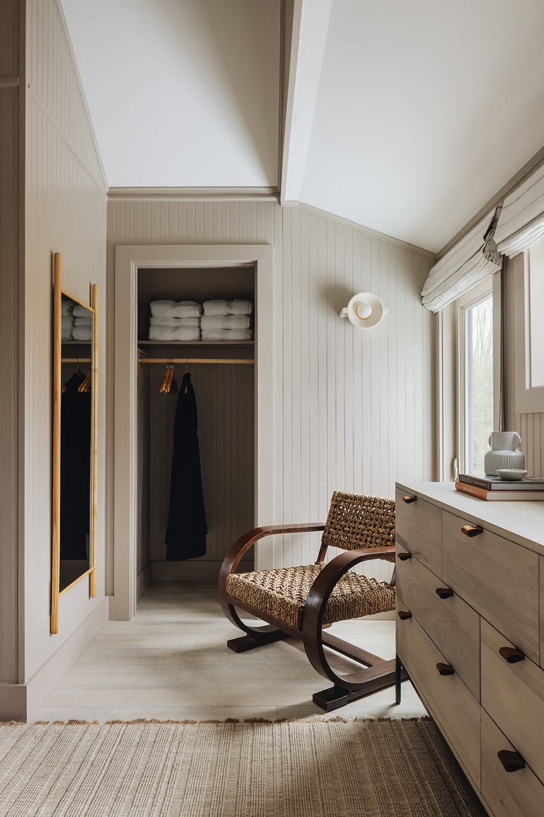 Serene bedroom with light wood paneling, a light-colored dresser with wooden knobs, a unique woven and wood accent chair, and a full-length mirror, leading to a closet.