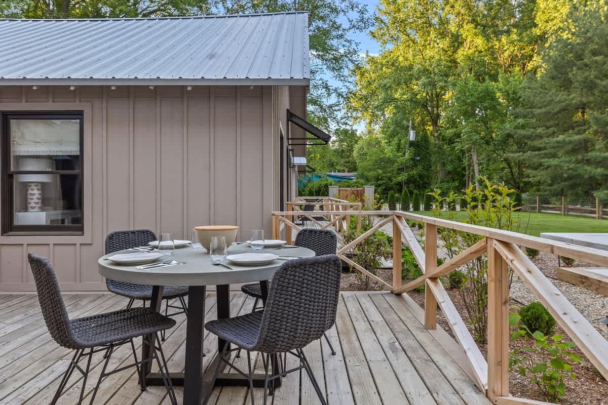 Outdoor wooden deck featuring a round dining table with four woven chairs, set against a backdrop of lush green trees and a glimpse of a pool area in the distance.