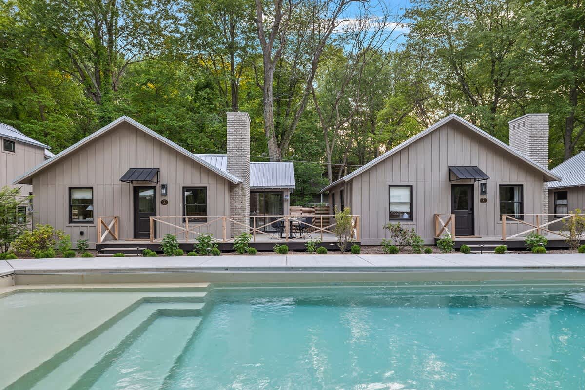 View of several modern farmhouse-style cottages from the edge of the swimming pool, showing their light-colored vertical siding, metal roofs, and individual small decks with railings. The pool has integrated steps, and lush trees are in the background.