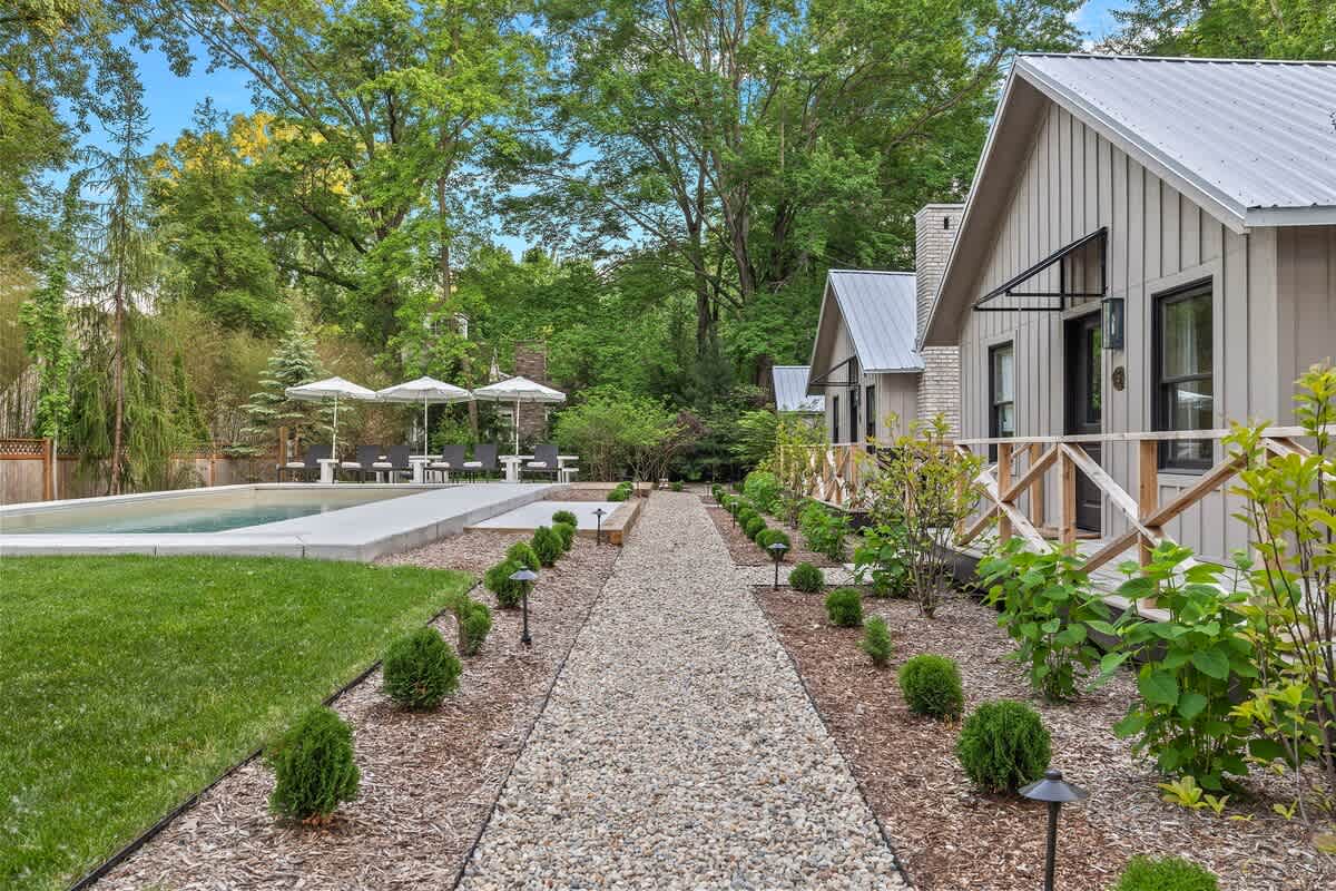 A wide view of a gravel pathway bordered by manicured shrubs and ground lights, running alongside several modern light-colored cottages with dark roofs and small porches, with a swimming pool area visible in the distance.