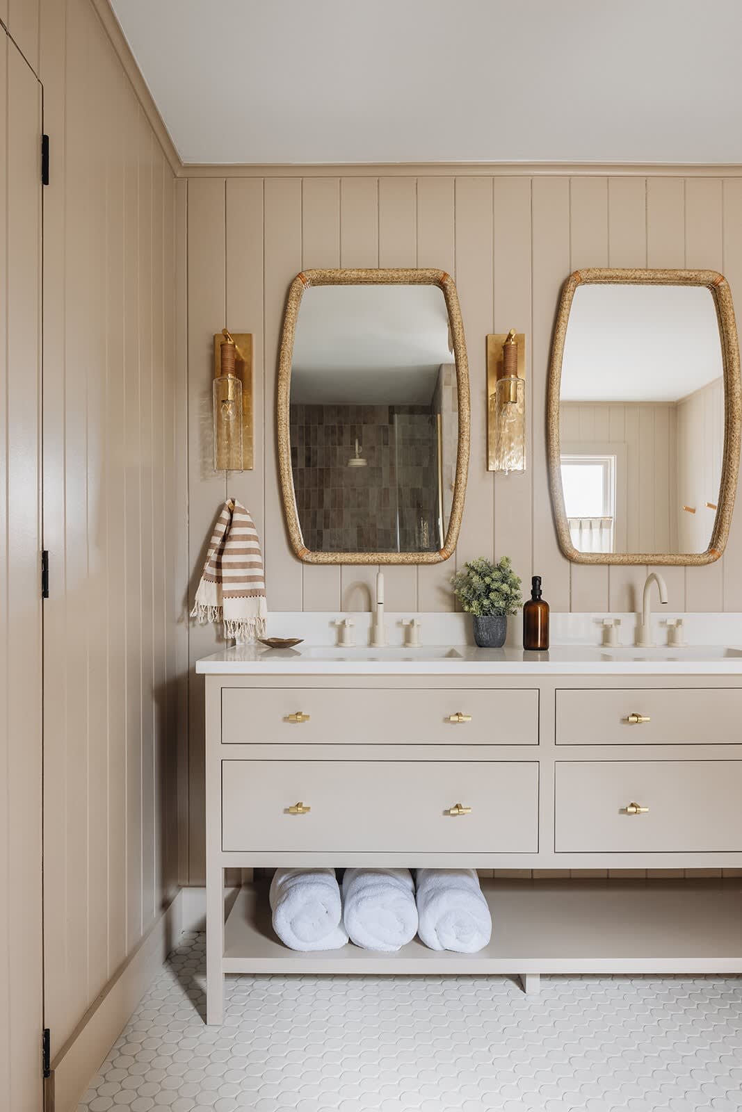 Stylish bathroom with a light beige double vanity featuring brass hardware, white countertop, and two woven mirrors with brass sconces. The walls are paneled in a warm neutral tone, and the floor is covered in white scallop-shaped tiles.
