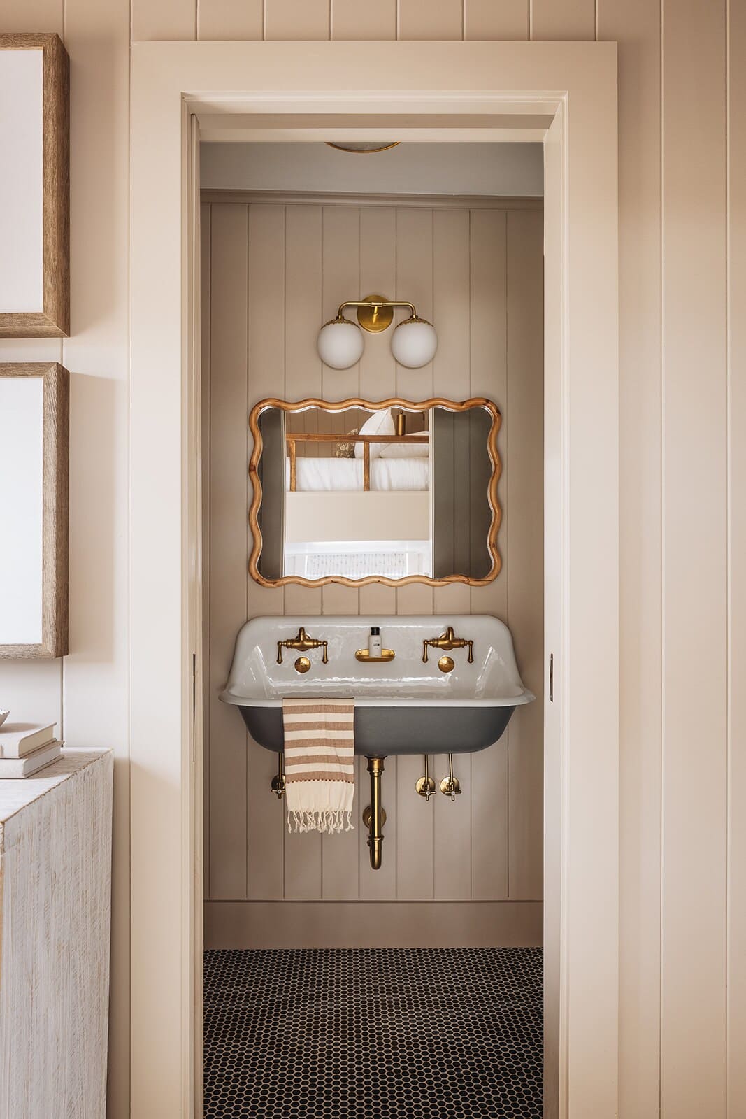A compact bathroom featuring a unique double basin sink with brass fixtures, a decorative mirror, and a globe light fixture, set against light wood paneling and a dark hexagonal tile floor.