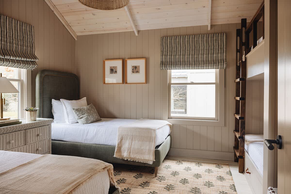 A cozy bedroom with two twin beds featuring green upholstered headboards and white bedding. The room has light brown vertical paneling walls, a vaulted ceiling, and a patterned rug, with a bunk bed structure visible on the right.
