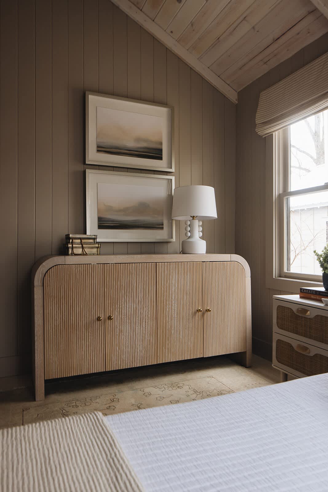 A light-filled bedroom corner featuring a natural wood fluted dresser with brass pulls, topped with a white lamp and framed artwork. The room has slatted wood walls and a vaulted wood-paneled ceiling.