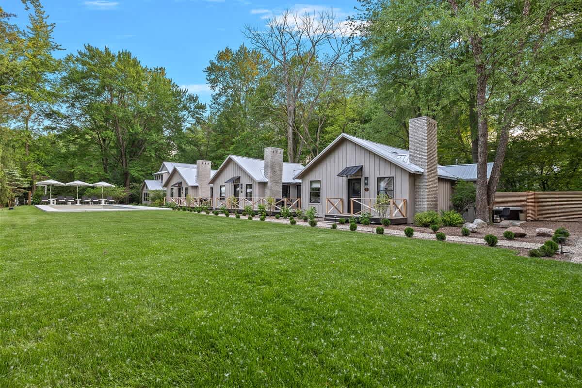 A panoramic view of a row of modern light-colored cottages, each with a dark roof, brick chimney, and small front porch, situated behind a vast, well-maintained green lawn, all framed by lush trees.