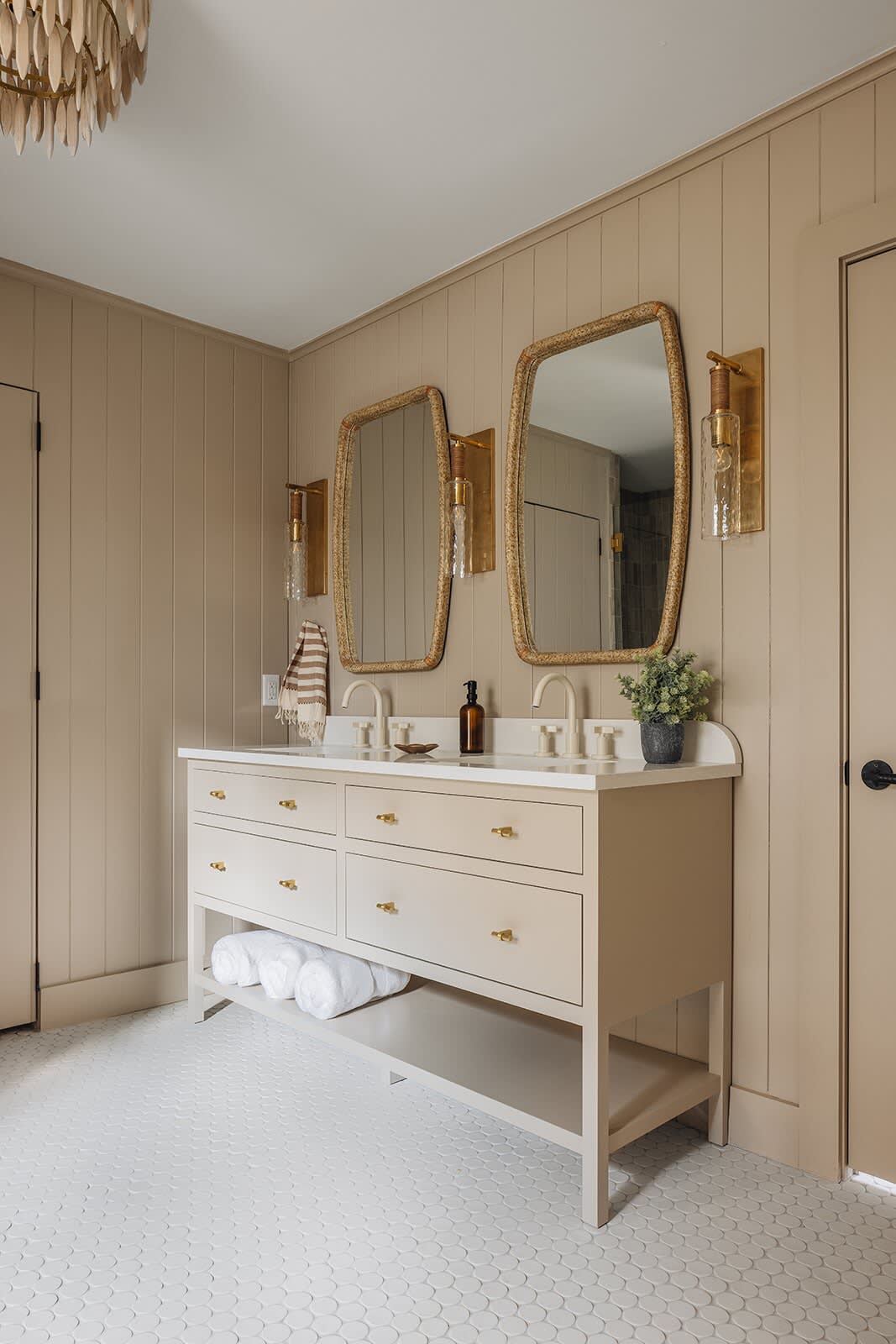 Elegant bathroom featuring a double vanity with a light beige cabinet, white countertop, and brass hardware, flanked by two woven mirrors and brass sconces. The walls are paneled in a warm beige, and the floor has white scallop-shaped tiles.