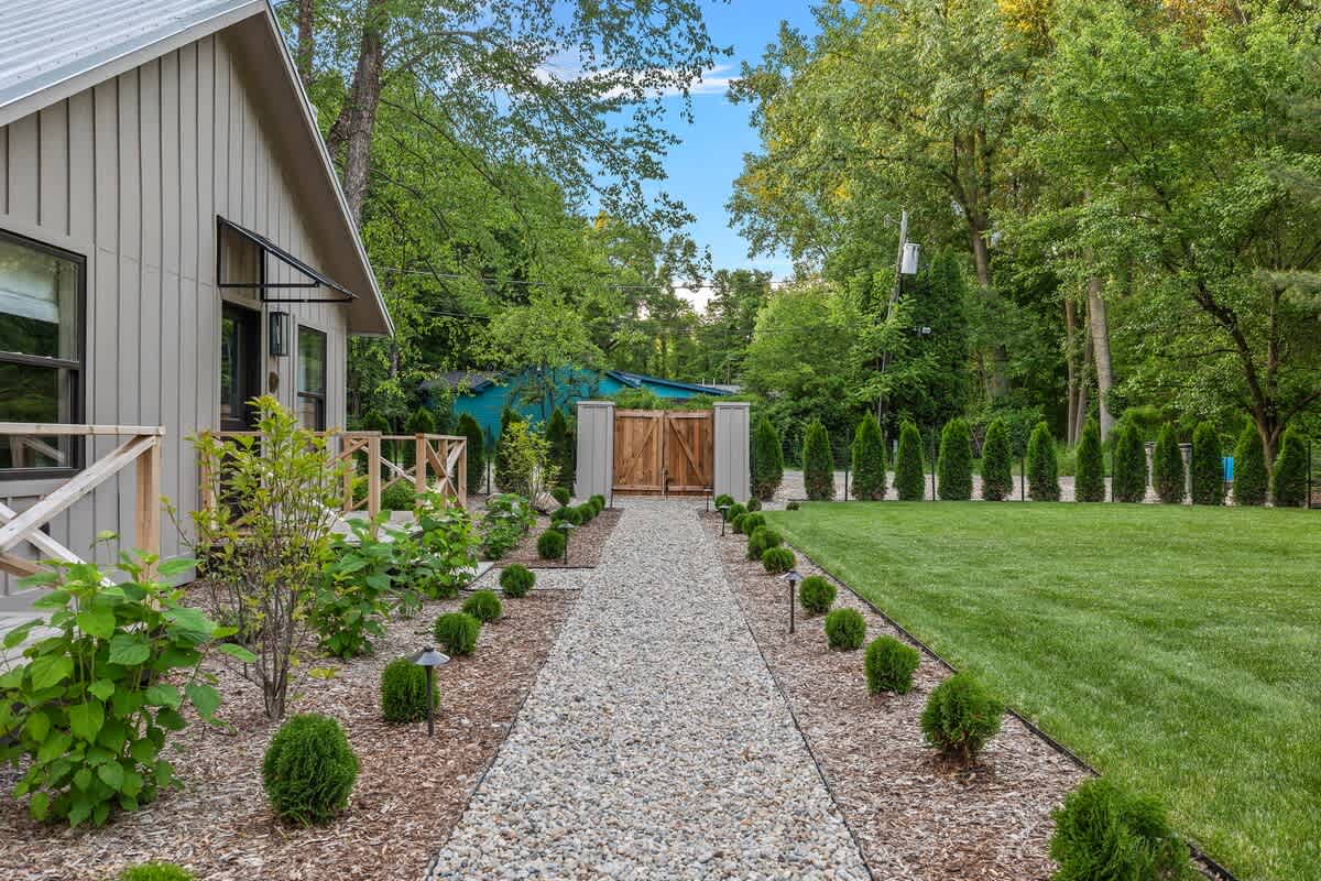 A neatly maintained gravel pathway lined with small green shrubs and ground lights, leading towards a wooden gate, with a modern cottage on the left and a vibrant green lawn on the right, all surrounded by tall trees.