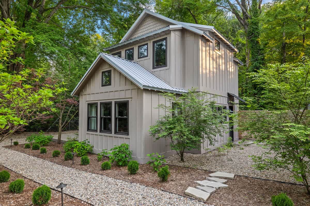 A charming two-story cottage with light gray board-and-batten siding, dark trim windows, and a metal roof, nestled among lush green trees. A gravel path with stepping stones leads to the entrance.