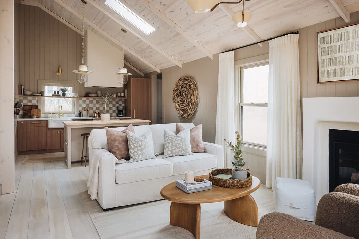 Bright and airy open-concept living room with a vaulted whitewashed wood ceiling, light wood floors, and a white sofa facing a wooden coffee table. The space connects to a kitchen area in the background.