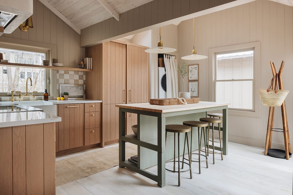 A bright kitchen with light wood cabinetry, a sage green island with bar stools, and a checkered tile backsplash, featuring a vaulted ceiling and light wood floors.