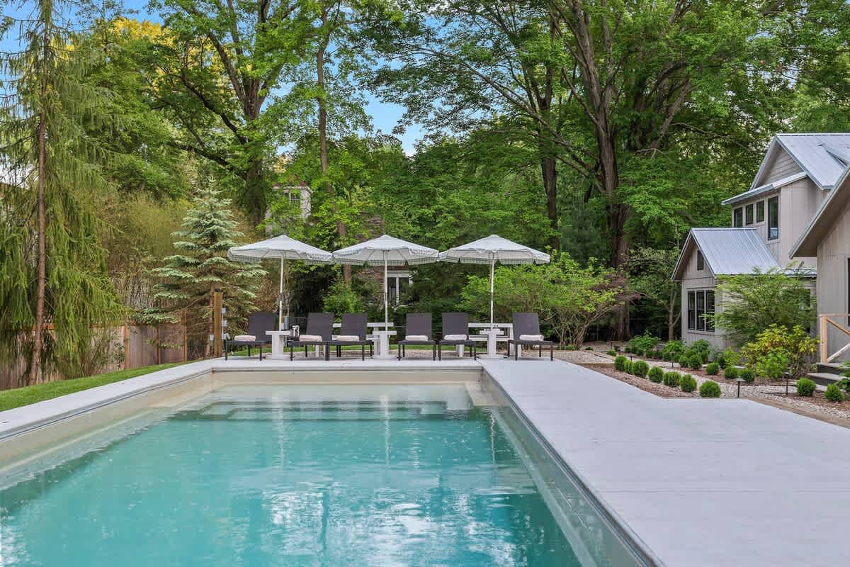 A long outdoor swimming pool with light blue water, surrounded by a concrete deck, leading to a lounging area with dark chairs and white umbrellas, with modern cottages and dense green foliage in the background.