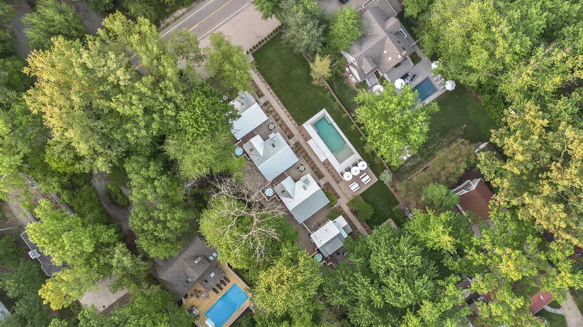 Overhead aerial view showcasing the property's layout with multiple cottages, a pool, and hot tubs, surrounded by a dense green tree canopy, adjacent to a road and other residential properties, providing a sense of its neighborhood context.