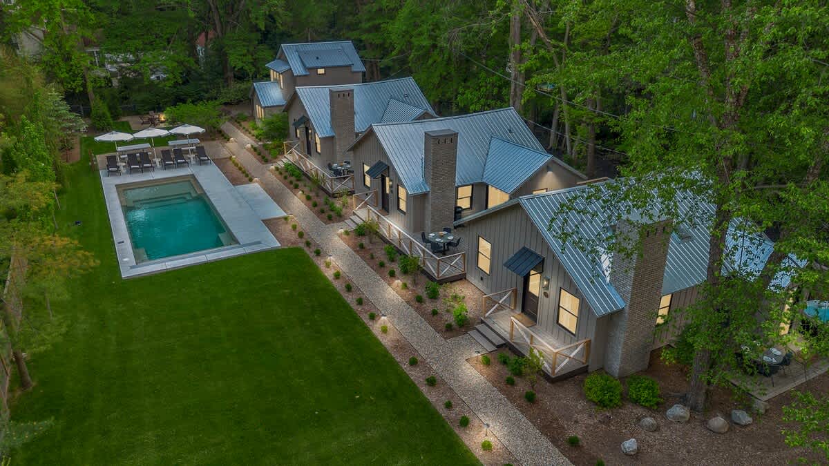 An aerial perspective showcasing the property's layout, with a row of modern cottages featuring dark roofs and chimneys, a gravel path, a rectangular swimming pool, and expansive green lawns, all nestled within a wooded area.