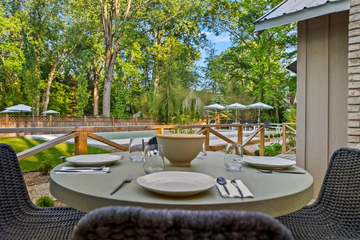 An outdoor dining table with place settings on a wooden deck, offering a view of a sparkling outdoor pool surrounded by lounge chairs and umbrellas. Lush green trees frame the background, creating a serene outdoor dining experience.
