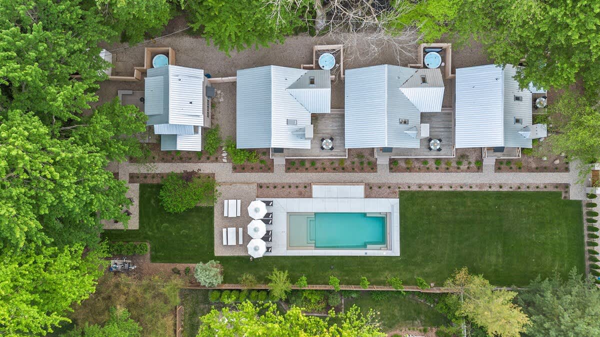 Top-down aerial view of the entire property, revealing multiple cottages with light metal roofs, several outdoor hot tubs, a rectangular swimming pool, and a large green lawn, all nestled within a dense canopy of green trees.