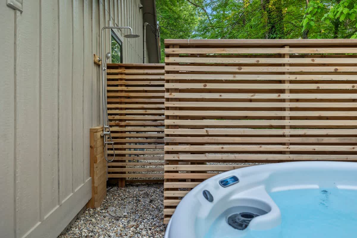 An outdoor shower with a chrome fixture is mounted on a light-colored siding wall, next to a slatted wooden privacy fence. A white hot tub with blue water is partially visible in the foreground, surrounded by gravel.