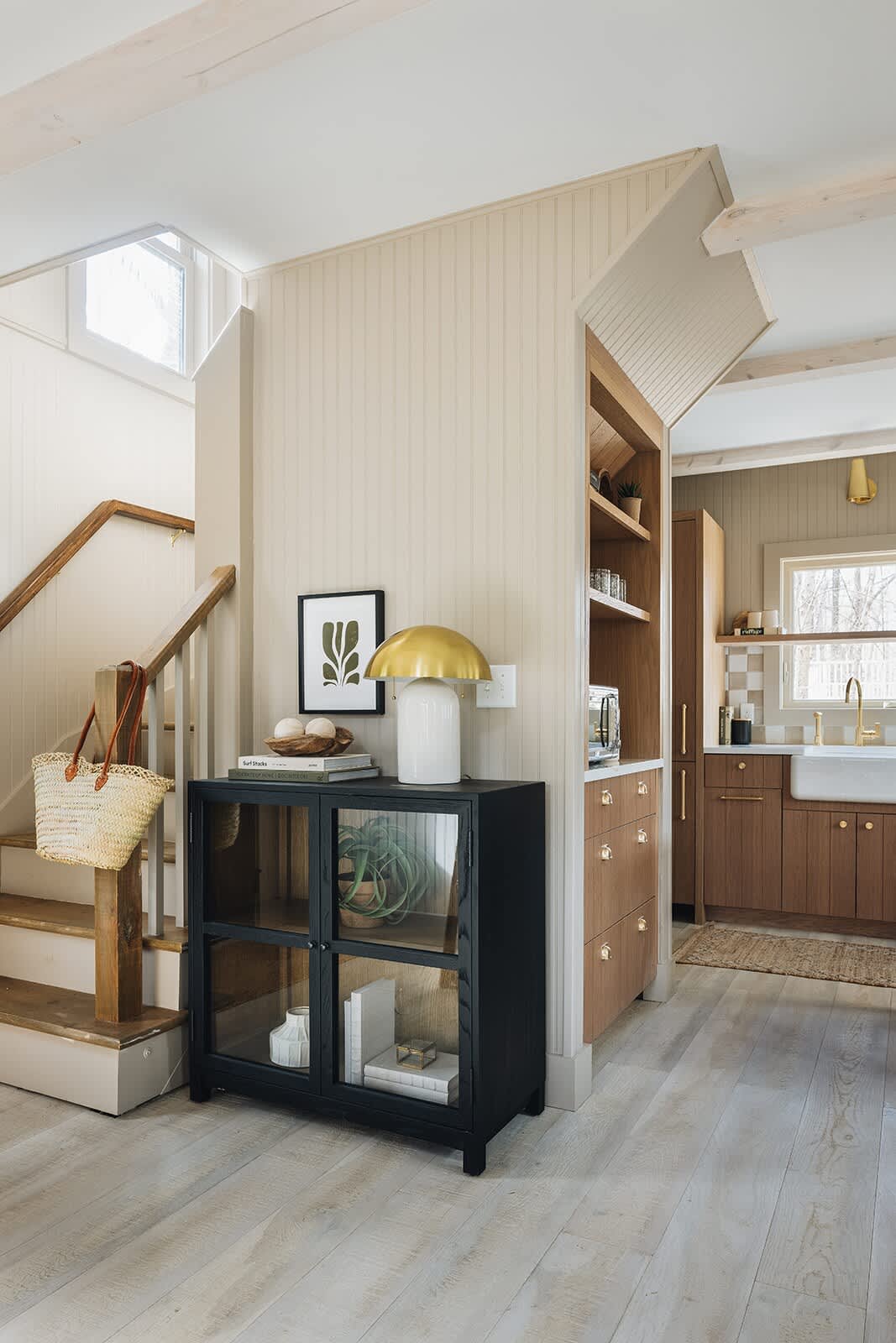 Inviting foyer area with light wood paneling and flooring, featuring a black glass-front cabinet, a gold mushroom lamp, and a staircase with a wooden handrail, leading towards the kitchen.
